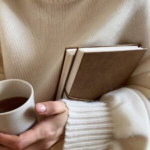 Woman holding books and a warm drink in a cosy knit jumper, the welcoming feel of a live wellbeing workshop