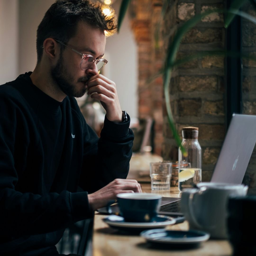 Man with glasses looking intently at laptop in a cafe, representing the pressure and mental load of always being on