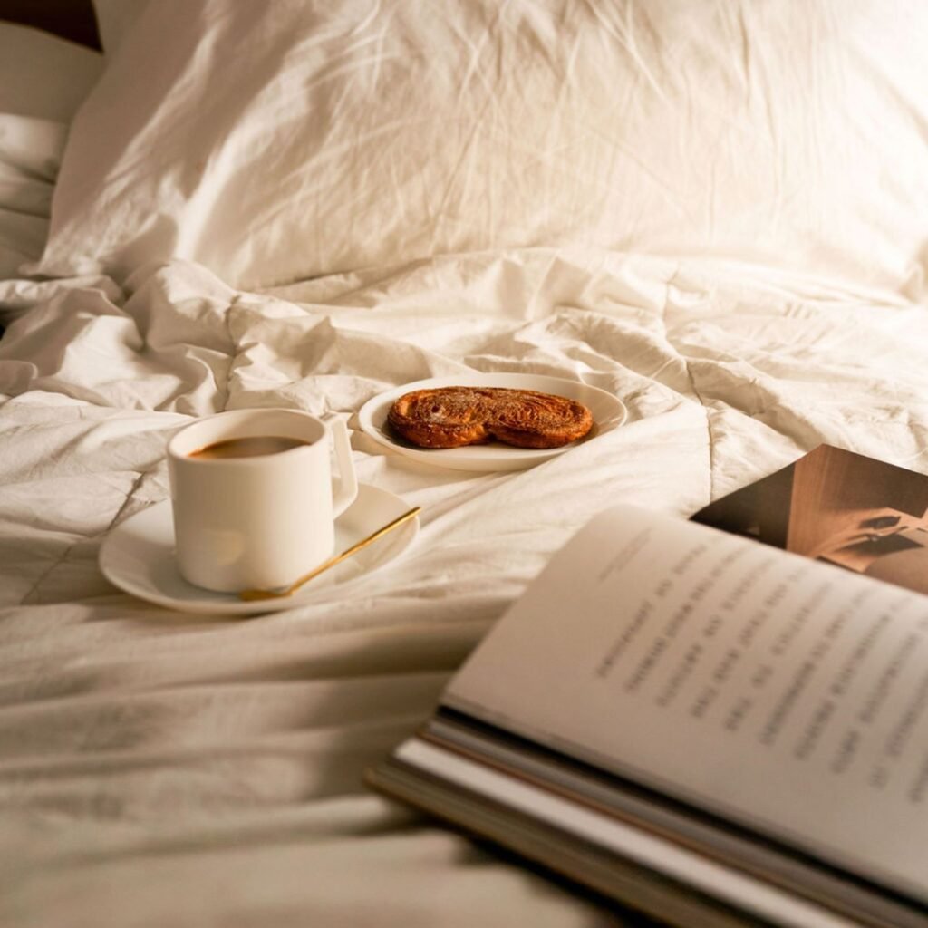 Morning coffee and pastry on a white saucer beside an open book on crumpled white bed linen, bathed in warm sunlight