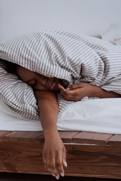 man with a striped duvet pulled over her head and one arm hanging out of bed, representing exhaustion and the struggle to face the day after poor sleep