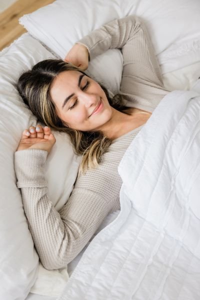 Woman lying in white bed with eyes closed and a relaxed smile, representing restful sleep after hypnotherapy for sleep and insomnia treatment