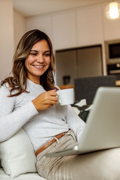 Woman sitting comfortably on a sofa with a coffee mug and laptop, smiling, representing the ease and flexibility of online ACT therapy sessions from home