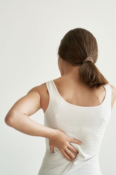 A woman from behind in a white vest top, hand resting on her lower back, representing chronic back pain