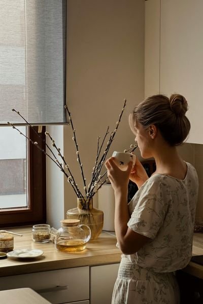 A woman in floral loungewear holding a mug by a kitchen window, an unhurried morning moment beside a vase of willow branches