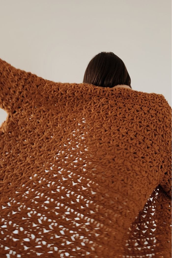 A person seen from behind, wrapped in a large rust-coloured crochet shawl, suggesting warmth, comfort and being held