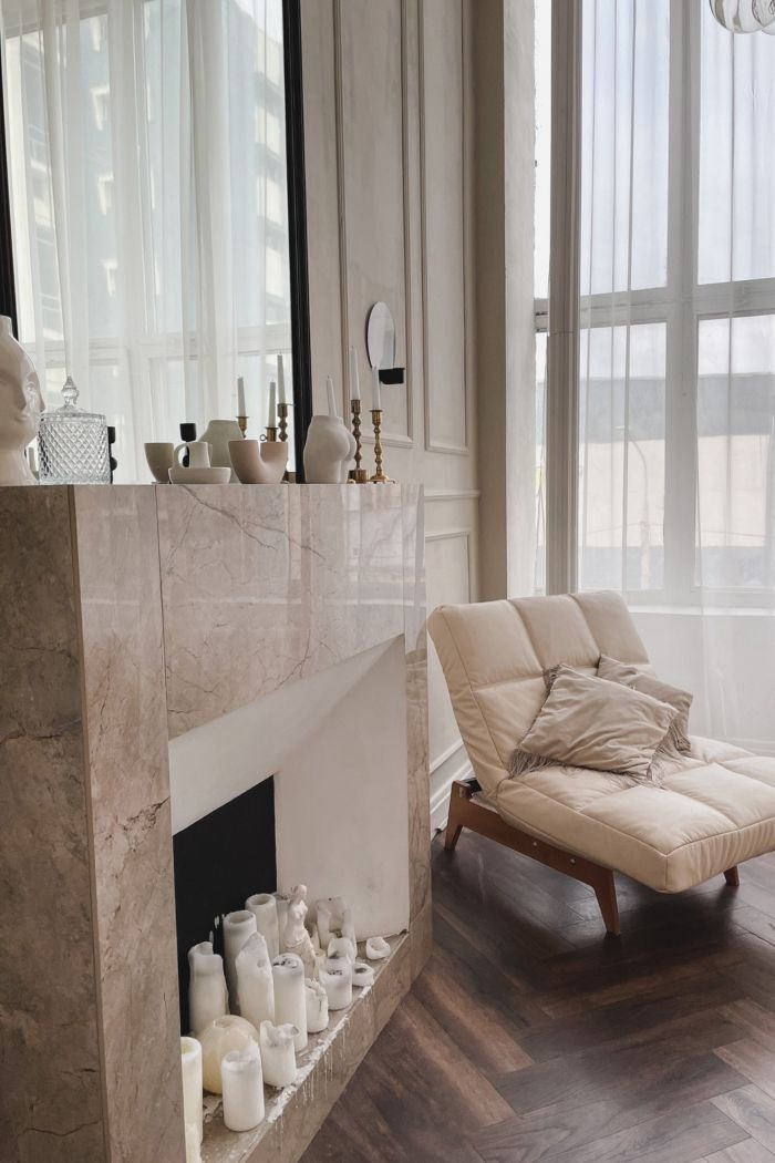 An elegant, light-filled room with a marble fireplace, cream lounge chair and tall sheer-curtained windows, evoking calm and spaciousness