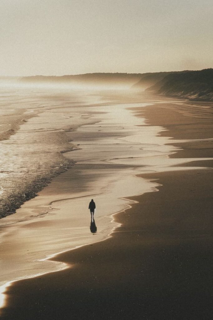 A lone figure walking along a vast sandy beach in warm, misty golden light, evoking space and perspective