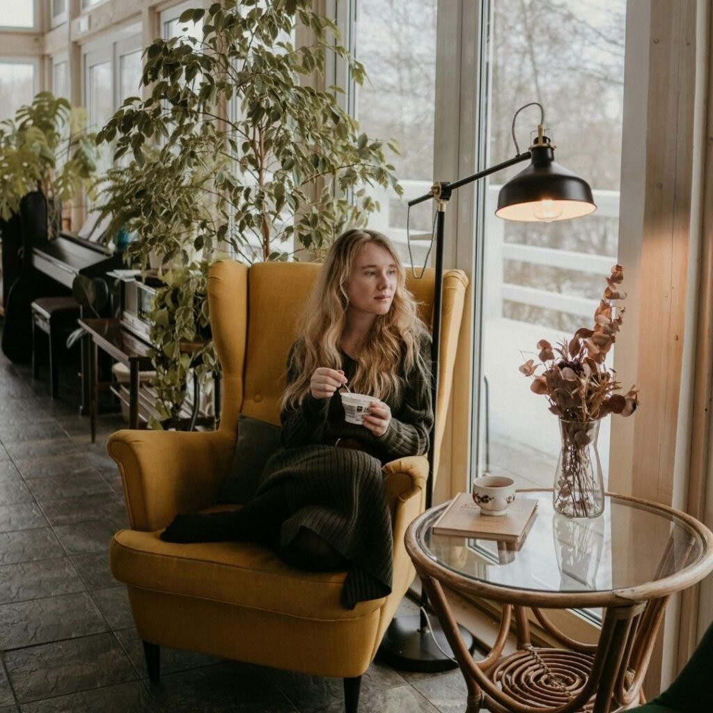 A woman sitting in a mustard yellow armchair in a plant-filled, warmly lit room, calm and present representing hypnotherapy for anxiety Warwickshire