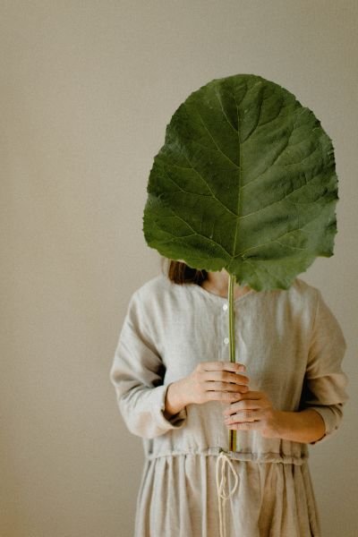 A woman partially concealed behind a large tropical leaf, capturing the instinct to hide away and withdraw that often comes with chronic pain