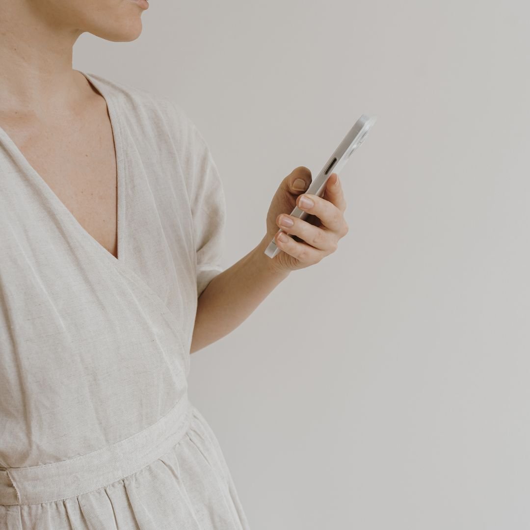 Woman in white holding a phone with the Your Mind Works app, representing the journalling and reflective practice that supports ACT work between sessions
