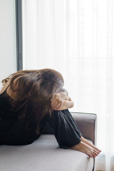 A woman in dark clothing hunched forward with her head in her hands on a light grey sofa, conveying the exhaustion of chronic pain