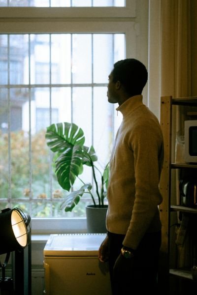 A man in a mustard yellow jumper standing in quiet profile by a window, a monstera plant behind him, lost in thought