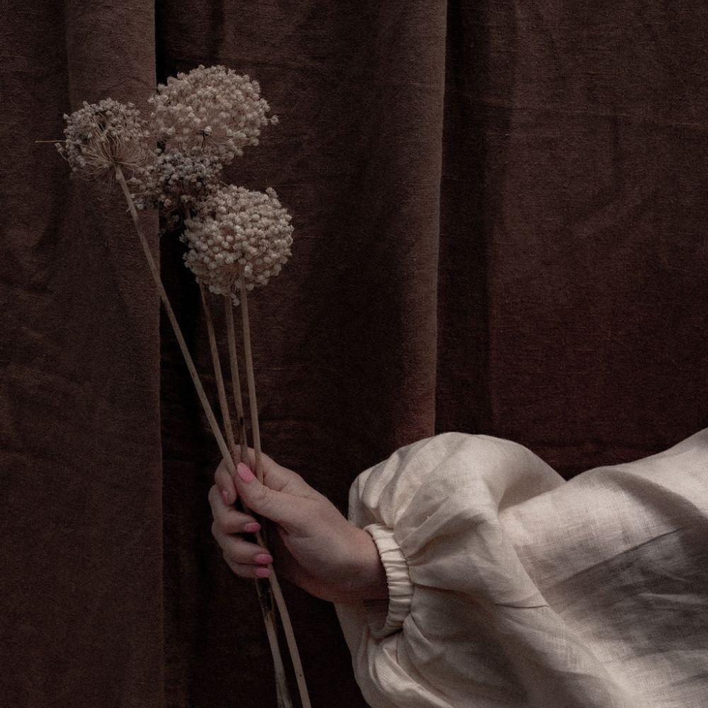 Woman in a linen shirt holding dried flowers against a dark curtain, a quiet and contemplative mood