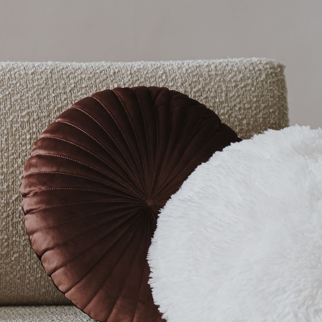 Two round cushions on a cream textured sofa, one deep brown velvet with a pleated fan detail, one soft white faux fur, representing the warm, tactile comfort of the therapy environment at Your Mind Works